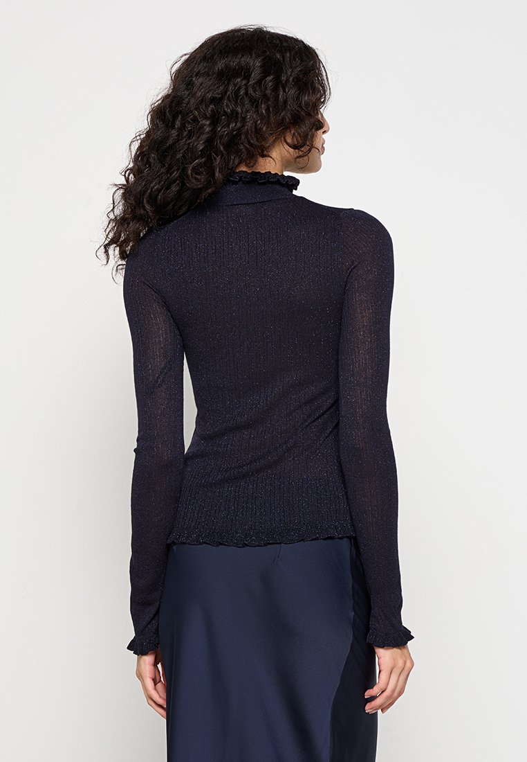 Haut à manches longues côtelé bleu marine avec un col montant, doté d'un ourlet volanté. Le matériau présente un léger éclat et une coupe près du corps.