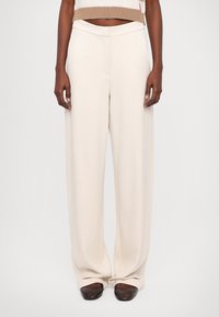 Beige weite Hosen mit glatter Textur, ausgestattet mit Seitentaschen und einem eng anliegenden Bund. Kombiniert mit dunklen, abgerundeten Schuhen.