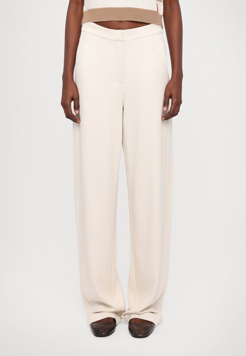 Beige weite Hosen mit glatter Textur, ausgestattet mit Seitentaschen und einem eng anliegenden Bund. Kombiniert mit dunklen, abgerundeten Schuhen.