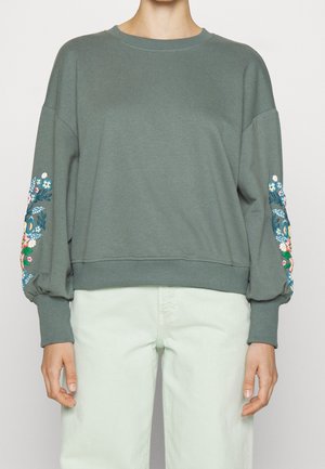 Osoba ubrana w luźną zieloną bluzę z haftem kwiatowym na bufiastych rękawach oraz jasne spodnie, stojąca na jednolitym tle.