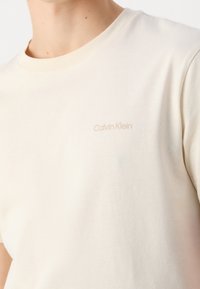 Camiseta de algodón de color beige claro con cuello redondo, que presenta un sutil logo de Calvin Klein en un tono ligeramente más oscuro en el pecho. Textura suave.