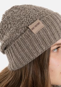 Bruine gebreide beanie met een gestructureerd patroon en een ribbelmanchet. Het heeft een leren label met "Knit Factory" erop geperst. Van zacht garen materiaal.