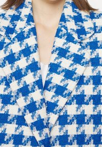 Gros plan d'une veste à motif pied-de-poule texturé bleu et blanc avec de larges revers portée sur un haut blanc.