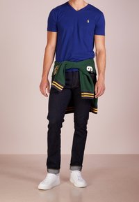 Royalblå T-shirt med V-ringning, mörk denimjeans, grön tröja knuten i midjan med gula accenter, och vita sneakers.