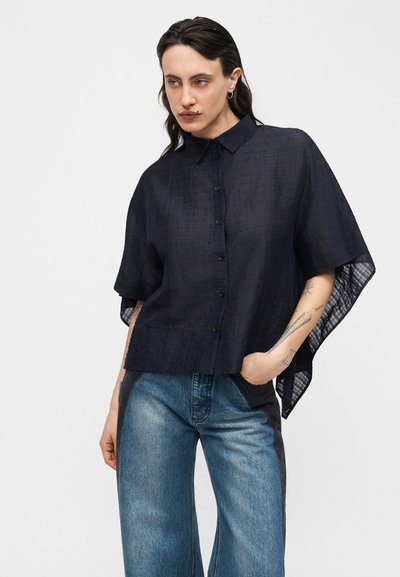 Άτομο με σκούρα μαλλιά, φοράει χαλαρό μαύρο πουκάμισο με κοντά μανίκια και κουμπιά και μπλε τζιν, με το ένα χέρι στην τσέπη, μπροστά σε απλό φόντο.