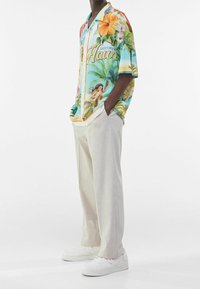 Short-sleeve button-up shirt with a colorful tropical print, featuring flowers, palm trees, and a vintage "Greetings from Hawaii" text. Paired with light beige trousers and white sneakers.