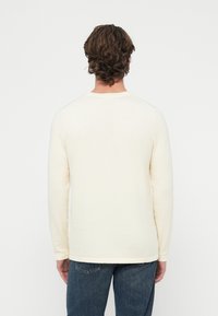 Jasnokremowa koszula z długim rękawem z okrągłym dekoltem i prążkowanym dołem, wykonana z miękkiego, teksturowanego materiału. Noszona z ciemnoniebieskimi dżinsami.