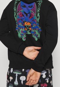 Schwarzer Hoodie mit lebhaftem Alien-Graphic und Pilzen, detaillierte Gestaltung in Blau, Lila und Grün. Begleitet von gemusterten Shorts.