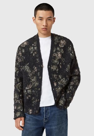 Cárdigan de patrón negro y gris con textura suave, que presenta un cierre de botones, cuello de chal y un ajuste ligeramente holgado. Lleva puesto sobre una camiseta blanca.