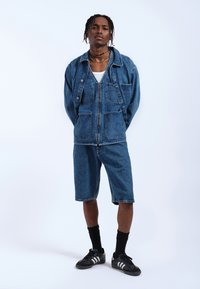 Chaqueta de mezclilla con múltiples bolsillos frontales y cierre de cremallera, combinada con pantalones cortos de mezclilla a juego. Zapatillas deportivas negras y calcetines oscuros completan el conjunto.