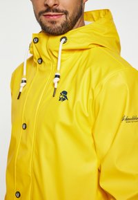 Gele, waterdichte regenjas met een capuchon, witte trekkoorden, zwarte drukknoopjes en een logo op de borst. Gladde, glanzende textuur.