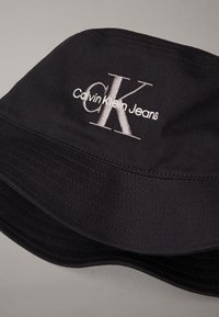 Black bucket hat with white embroidered "cK Calvin Klein Jeans" logo on the front, displayed against a plain gray background.