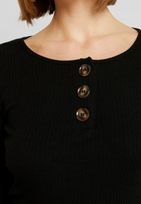 Zwart geribbeld langarmtop met een ronde halslijn en vier grote, glanzende bruine knopen in het middenvoor. Zachte textuur.