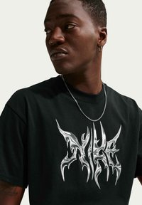 Νεαρός άνδρας που φοράει μαύρο γραφικό T-shirt με ασημένιο αφηρημένο σχέδιο, ασημένιο κολιέ αλυσίδα και κρίκο σκουλαρίκι, κοιτάζοντας προς τα αριστερά σε απλό φόντο.