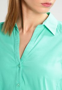 Camicia verde menta con colletto e bottoniera, caratterizzata da una texture liscia e una patta frontale. I bottoni sono visibili lungo il centro.