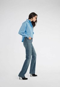 Camicetta azzurra corta con maniche lunghe, abbinata a jeans svasati azzurri sbiaditi e scarpe con tacco nere. La modella posa di profilo.