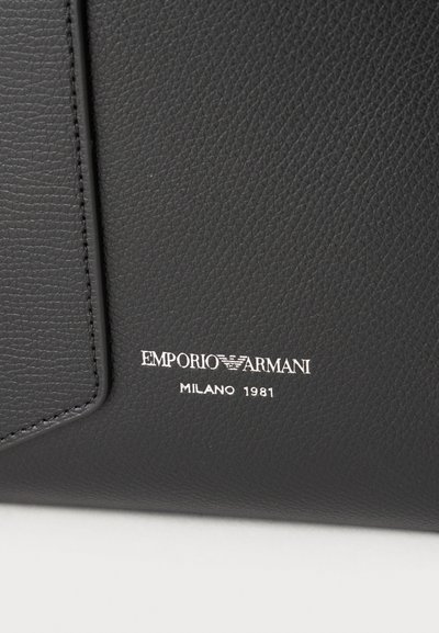 Μαύρη υφή δερμάτινης επιφάνειας με λευκό λογότυπο Emporio Armani Milano 1981 ανάγλυφα κοντά στην ραμμένη άκρη.