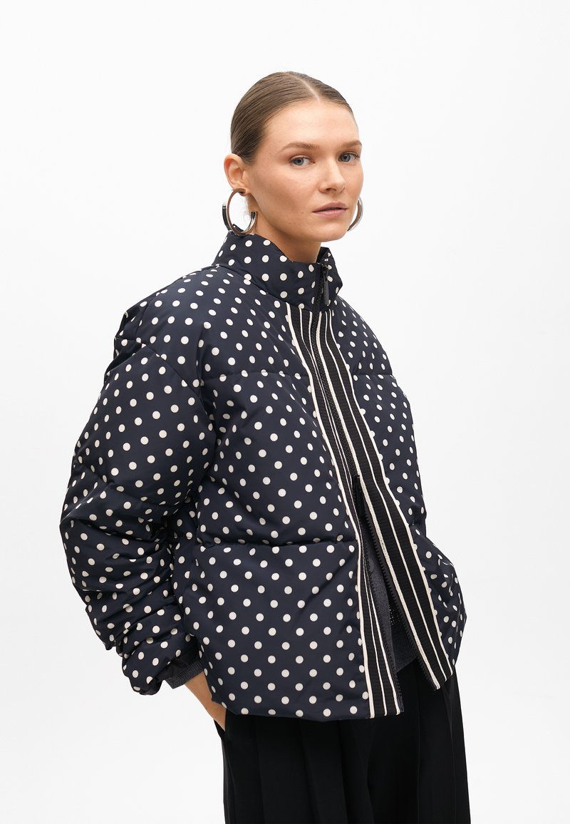 Námořnická pufrová bunda s puntíky, vysokým límcem, svislým zipem a texturovanou tkaninou; má oversized siluetu a boční kapsy.