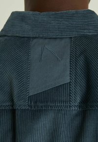 Blue corduroy jacket collar with textured fabric, featuring a contrasting smooth patch and a stitched detail resembling an abstract letter.