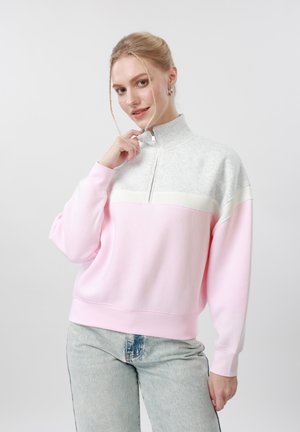 Frau mit blonden Haaren, die einen rosa-grauen Sweatshirt mit Viertelreißverschluss und hellblaue Jeans trägt, posiert vor einem schlichten grauen Hintergrund.