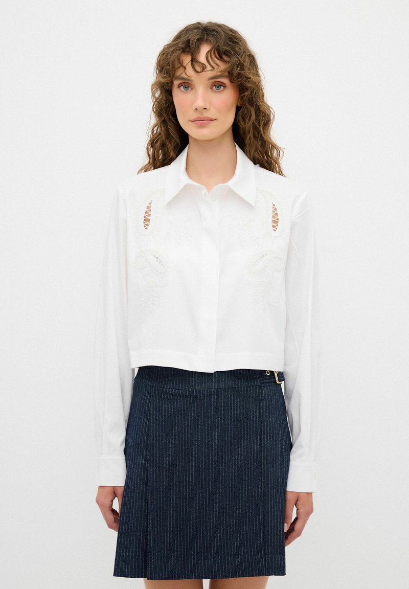Weiße, verkürzte Bluse mit langen Ärmeln, die mit gestickten Details und Ausschnitten versehen ist. Kombiniert mit einem dunkelblauen Streifenrock.