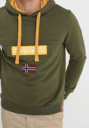 Mand iført olivengrøn hættetrøje med gule snore, Napapijri-logo lap og norsk flag-lap på brystet, beige bukser.