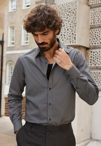 Chemise à boutons en gris foncé avec un motif circulaire, faite de tissu tissé. Elle possède des manches longues, un col et une fermeture à boutons sur le devant.