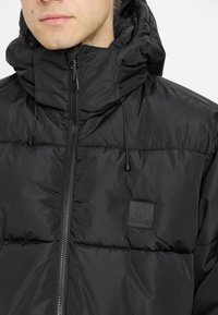 Veste rembourrée noire avec un col haut et une capuche amovible. Comprend une fermeture éclair sur le devant, des cordons de serrage réglables et un écusson de marque sur la poitrine.