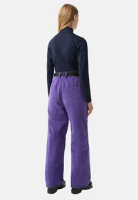 Žena stojící zády, oblečená v tmavě modrém dlouhém rukávu, fialových manšestrových kalhotách, černém opasku a černých botách na bílém pozadí.