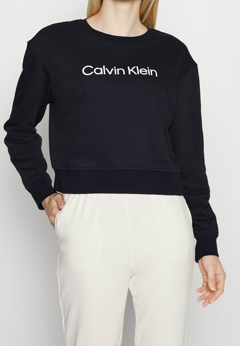Femme portant un sweat-shirt Calvin Klein noir avec un logo blanc et un pantalon blanc, main dans la poche sur un fond clair uni.