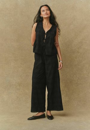 Haut sans manches en coton noir avec col en V, bordure en dentelle et cordon de serrage. Associé à un pantalon noir à jambes larges avec un motif texturé.