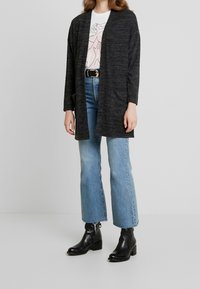 Grå stickad kavaj med öppen framsida, långa ärmar, kombinerad med en grafisk vit skjorta, ljusblå utsvängda jeans och svarta ankelstövlar.