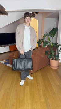 Sort læder duffel taske med to top håndtag, glat tekstur og et stort hovedrum. Sat mod en moderne indendørs baggrund.