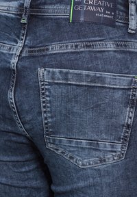 Jeans de mezclilla azul oscuro con un bolsillo trasero, costuras visibles y una etiqueta de marca situada por encima de la cinturilla. Tejido texturizado.
