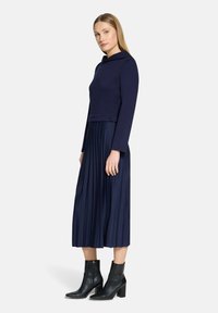 Granatowy dwuczęściowy strój składający się z dopasowanego topu z kapturem oraz plisowanej spódnicy midi, zestawiony z czarnymi botkami na obcasie.