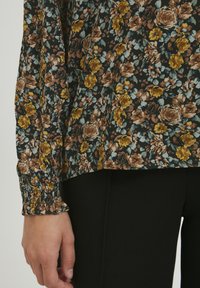 Floralbluse mit schwarzem Hintergrund und lebhaften gelben und braunen Rosen, verziert mit gesmokten Bündchen und einem lockeren Schnitt.