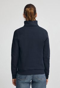 Felpa blu navy con colletto alto, maniche lunghe e orlo a coste. Realizzata in tessuto morbido, presenta una texture liscia e un design minimale.