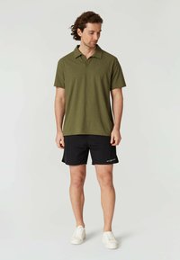 Olivgrön polo-shirt med krage, kombinerad med svarta shorts. Shorts har en logotyp på sidan och kompletteras av vita sneakers.