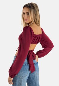 Bordeauxrotes, langärmeliges Crop-Top mit quadratischem Ausschnitt, Puffärmeln, offenem Rücken und Bindetail. Kombiniert mit hochgeschnittenen blauen Jeans.