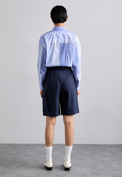 Tenue masculine comprenant une chemise rayée bleu clair et blanche, un short à rayures navy, et des chaussettes blanches avec des chaussures à talons blocs crème.