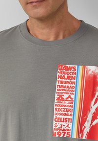 Graues Baumwoll-T-Shirt mit einem grafischen Aufnäher, der rote und weiße Buchstaben, Streifen in Blau und Gelb sowie einen abstrakten roten Hintergrund zeigt.