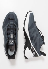 Donkergrijze hardloopschoenen met een gestructureerde bovenkant, zwarte veters en witte accenten. Beschikt over een robuuste zool met een opvallend profiel.