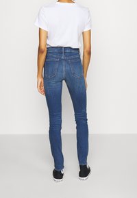 Jeans ajustados de tiro alto en azul con cinco bolsillos, costura sutil y una textura suave, combinados con zapatillas negras y una camiseta blanca.