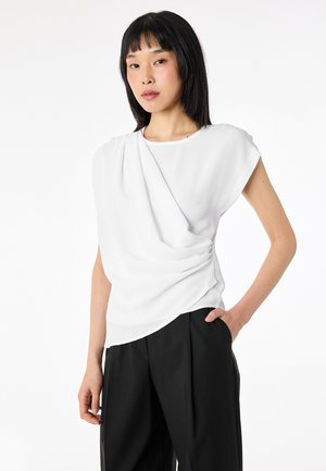 Giovane donna con capelli neri che indossa un top bianco drappeggiato senza maniche e pantaloni neri a gamba larga, in piedi con una mano in tasca.