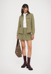 Alyvuogių žalia tweed striukė su rudi mygtukais, atitinkantys sijonas, balta marškiniai, leopardų rašto rankinė ir blizgūs burgundiški iki kelių ilgio batai.