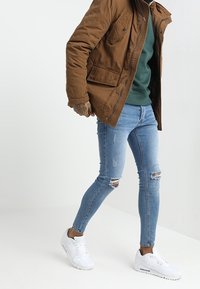 Bruine canvas jas, groene trui, versleten lichtblauwe skinny jeans en witte sneakers. Kenmerken zijn scheuren in de jeans en zakken van de jas.