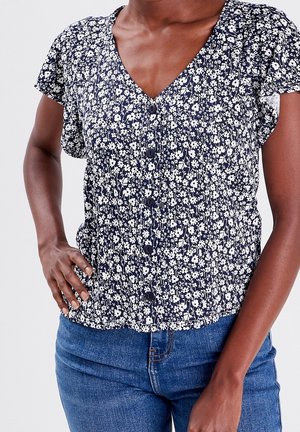 Blouse bleu marine à motifs floraux avec un col en V, des manches courtes évasées, une patte de boutonnage avant et un ourlet élastique au bas. Présente des motifs de fleurs blanches sur toute la surface.