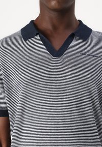 Prúžkovaná polo košela v námorníckej modrej a bielej farbe, s výstrihom do V, krátkymi rukávmi a tmavým golierom. Tkanina sa zdá byť hladká s jemným leskom.