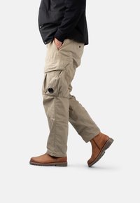 Pantalones cargo beige con múltiples bolsillos, detalle de botón en el lado y un corte relajado. Combinados con botas de tobillo marrones.