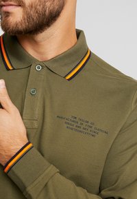 Oliwkowa koszulka polo z dwiema guzikami, z kołnierzykiem w pomarańczowe i granatowe paski. Niebieski napis z logo na klatce piersiowej.